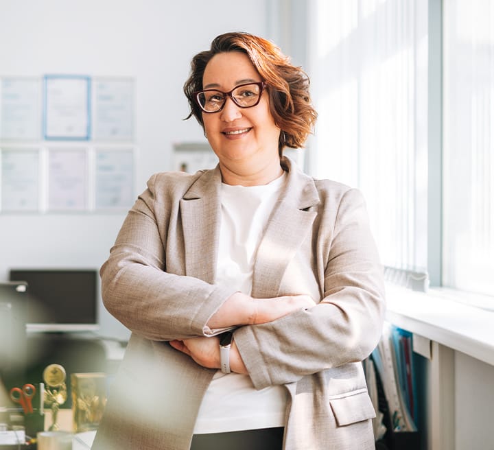Eine Frau, die professionell gekleidet ist, verschränkt die Arme vor der Brust und schaut lächelnd in die Kamera.
