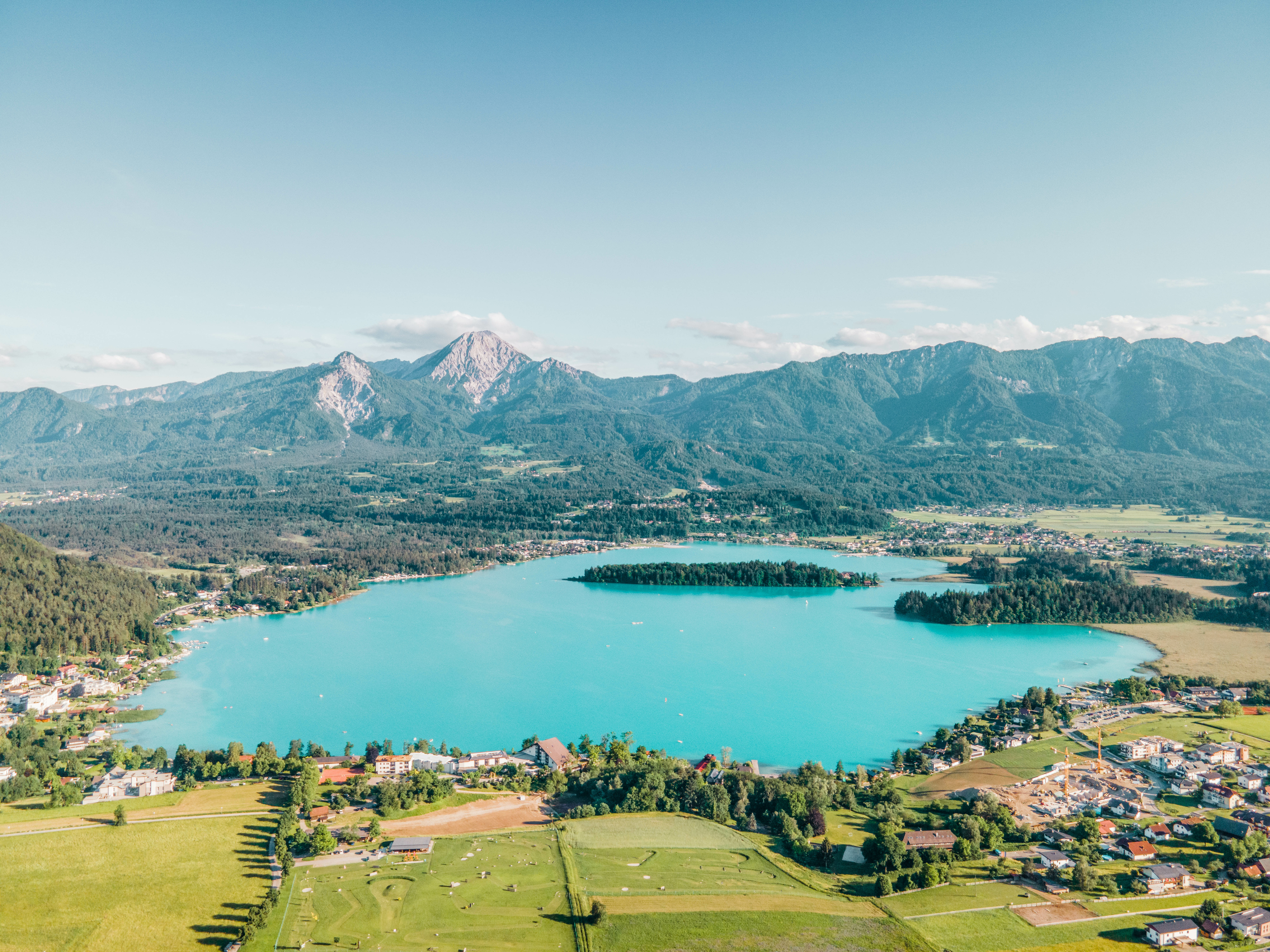 Bäume und Felder befinden sich um den strahlend blauen Faaker See. In der Mitte des Sees ist eine kleine Insel.