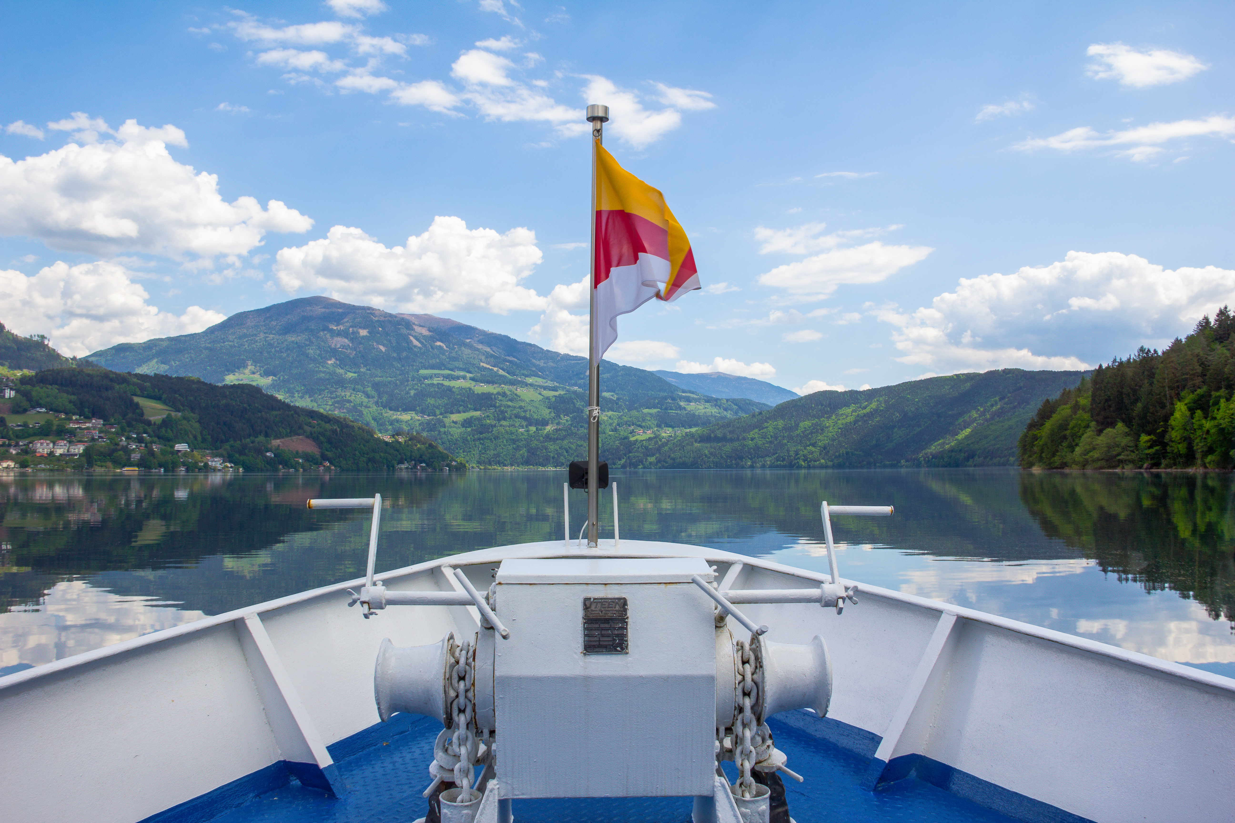 Die Spitze eines Bootes, auf dem eine Fahne steht, ragt in die Höhe. Das Bild wurde vom Boot aus gemacht und rundherum befinden sich Berge und ein See. 