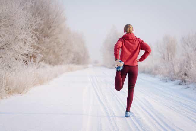 Eine Frau steht mit Sportkleidung auf einem Feldweg, der mit Schnee bedeckt ist. Sie hat ihr Bein nach hinten weggestreckt und hält ihren Fuß in ihrer Hand.
