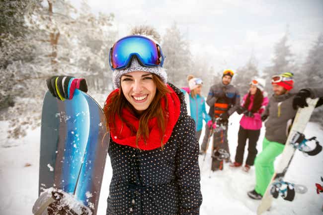 Eine Gruppe von Leuten mit Skianzügen und Snowboards unterhält sich in einer verschneiten Landschaft. Eine Person steht weiter vorne und schaut lächelnd in die Kamera.