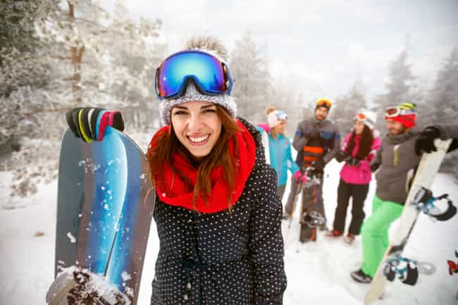 Eine Gruppe von Leuten mit Skianzügen und Snowboards unterhält sich in einer verschneiten Landschaft. Eine Person steht weiter vorne und schaut lächelnd in die Kamera.