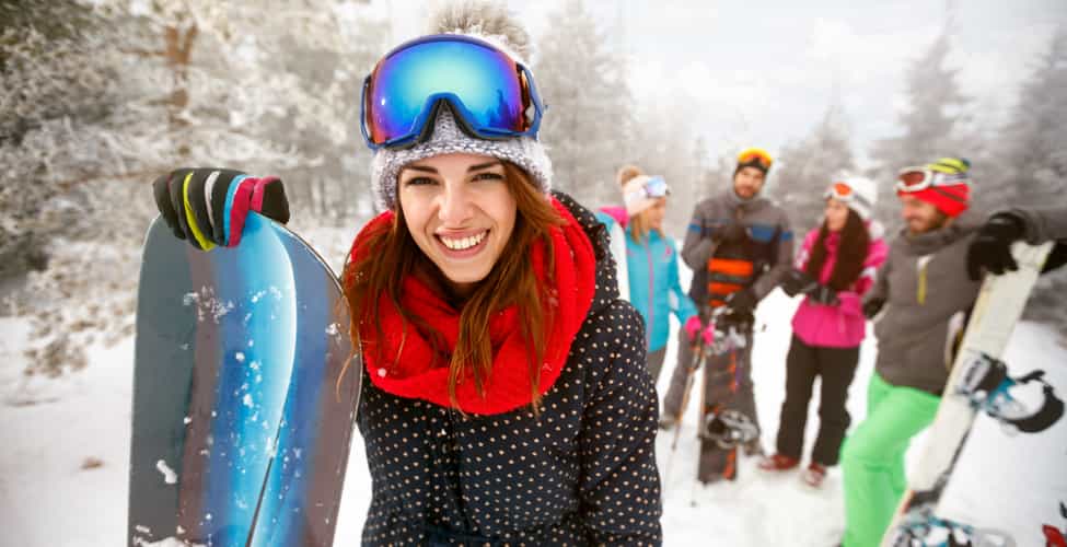Eine Gruppe von Leuten mit Skianzügen und Snowboards unterhält sich in einer verschneiten Landschaft. Eine Person steht weiter vorne und schaut lächelnd in die Kamera.