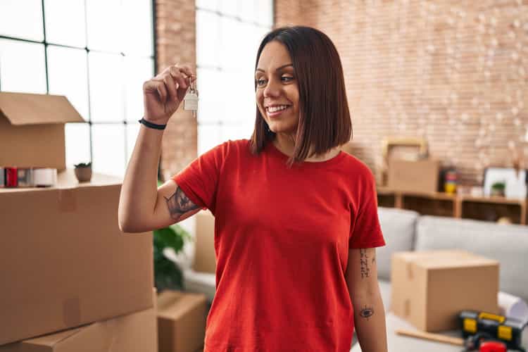 Eine Frau mit rotem Shirt steht in der Mitte eines Raumes und hält lächelnd einen Schlüssel in der Hand. Um sie herum befinden sich Umzugskartons.