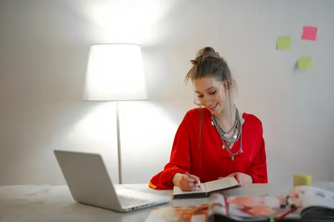 Junge Frau in roter Bluse schreibt lächelnd in ein Notizbuch an einem Schreibtisch mit Laptop und Zeitschriften.