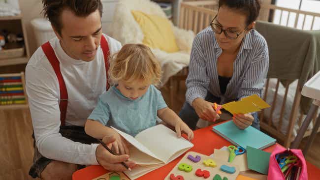 Eltern basteln mit ihrem kleinen Kind an einem Tisch. Der Vater hilft beim Malen, die Mutter schneidet Papier aus.