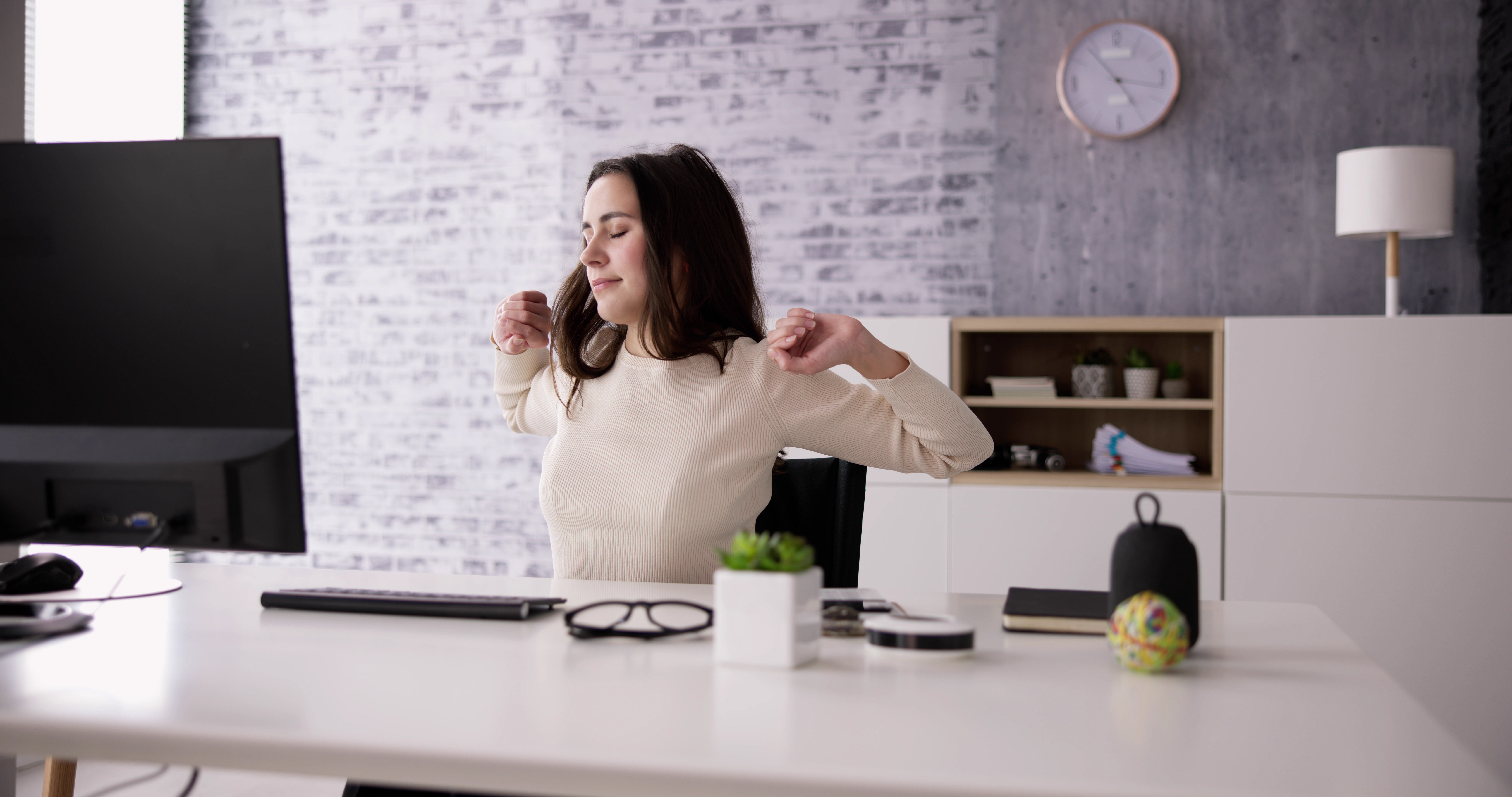Eine junge Frau mit braunen Haaren sitzt an ihrem Schreibtisch. Sie dehnt sich, indem sie ihre Arme seitlich hoch streckt. 