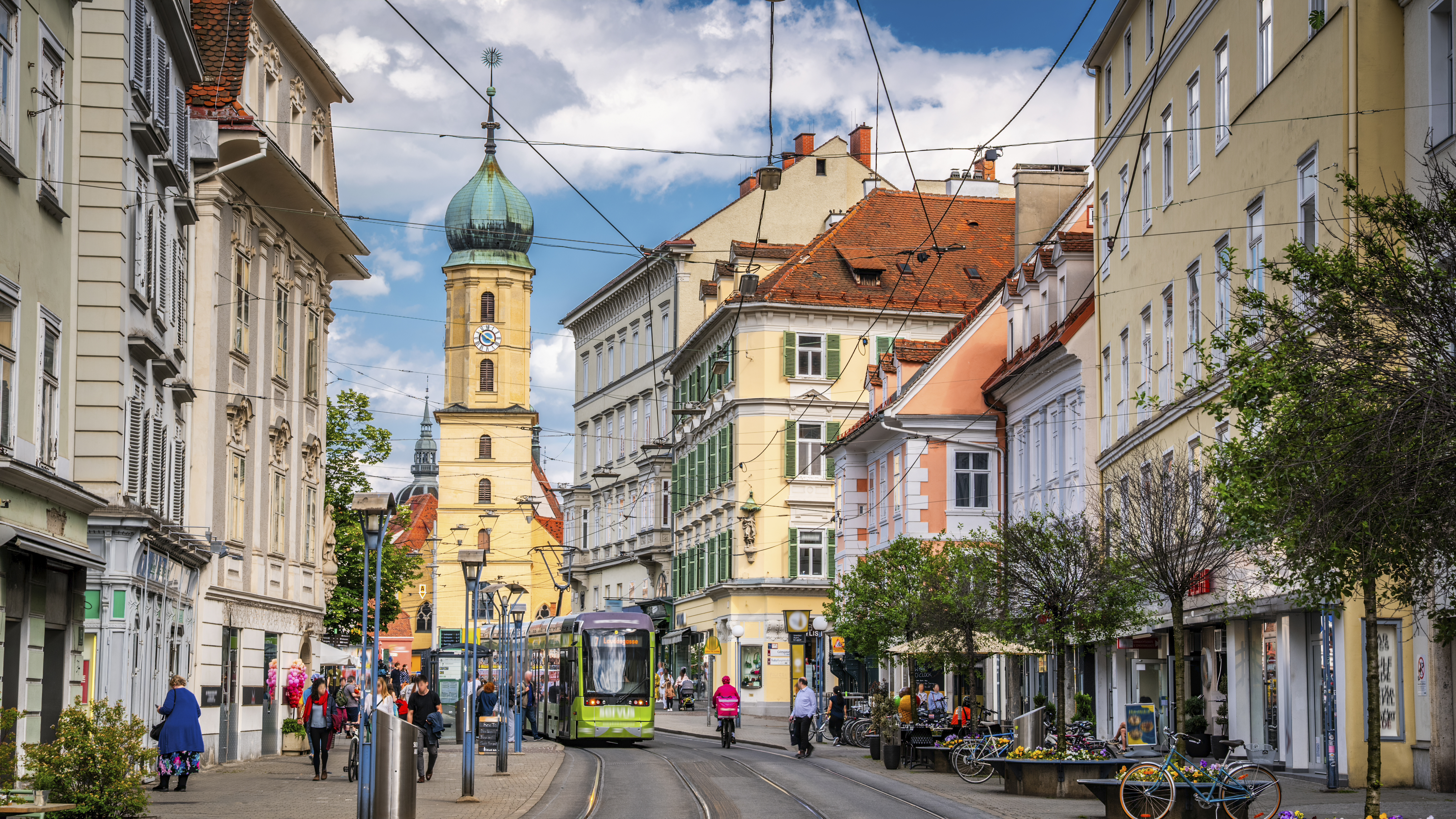 Street scene in Graz, Austria. A green tram passes through a lively avenue lined with pastel-colored buildings, shops, and pedestrians under a bright blue summer sky. The historic church tower of Mariahilferkirche (Church of the Mariahilf) in the background adds to the old-town character of this picturesque Styrian capital. Graz capital of the state of Styria in Austria - Europe