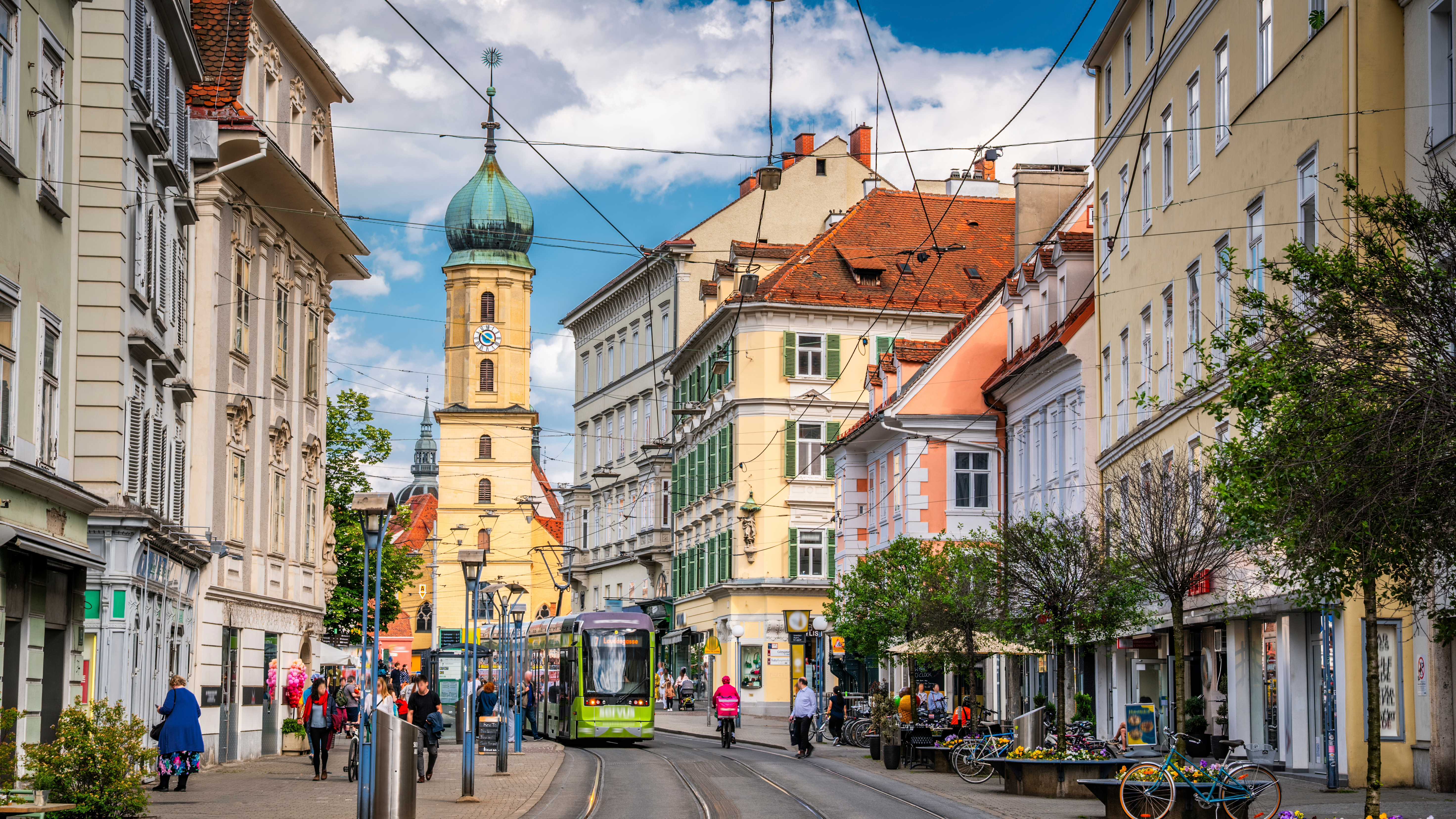 Street scene in Graz, Austria. A green tram passes through a lively avenue lined with pastel-colored buildings, shops, and pedestrians under a bright blue summer sky. The historic church tower of Mariahilferkirche (Church of the Mariahilf) in the background adds to the old-town character of this picturesque Styrian capital. Graz capital of the state of Styria in Austria - Europe