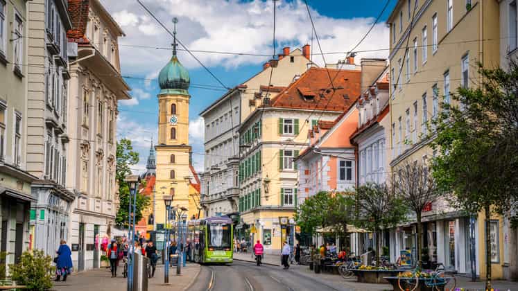 Street scene in Graz, Austria. A green tram passes through a lively avenue lined with pastel-colored buildings, shops, and pedestrians under a bright blue summer sky. The historic church tower of Mariahilferkirche (Church of the Mariahilf) in the background adds to the old-town character of this picturesque Styrian capital. Graz capital of the state of Styria in Austria - Europe