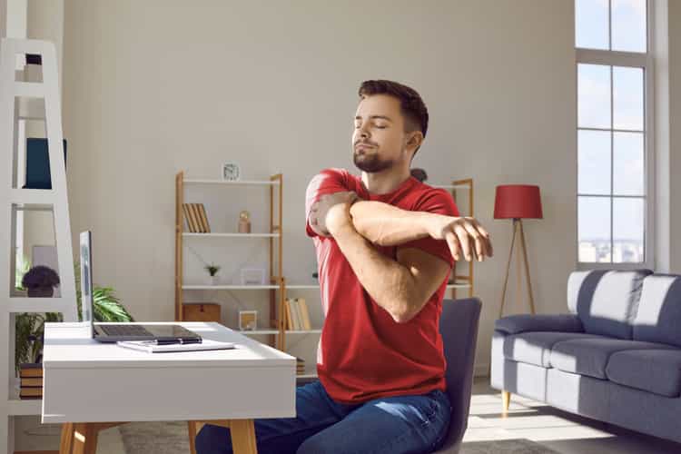 Ein Mann sitzt beim Schreibtisch und dehnt sich. Er streckt seinen rechten Arm in die linke Richtung und drückt mit seinem linken Arm seinen rechten Oberarm zu sich.