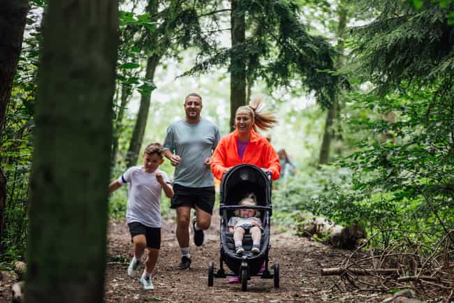 A wide shot of a group of adults and children wearing sports clothing. They are taking part in a fun run with other children and adults at Cramlington Nature Reserve in the North East of England. A woman runs through a forest with other participants as she pushes her daughter in a baby stroller.