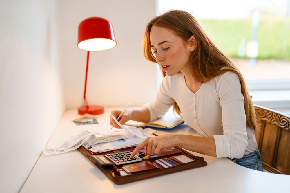 Junge Frau mit langen roten Haaren sitzt am Schreibtisch, rechnet mit Taschenrechner und prüft Belege bei Schreibtischlampe.