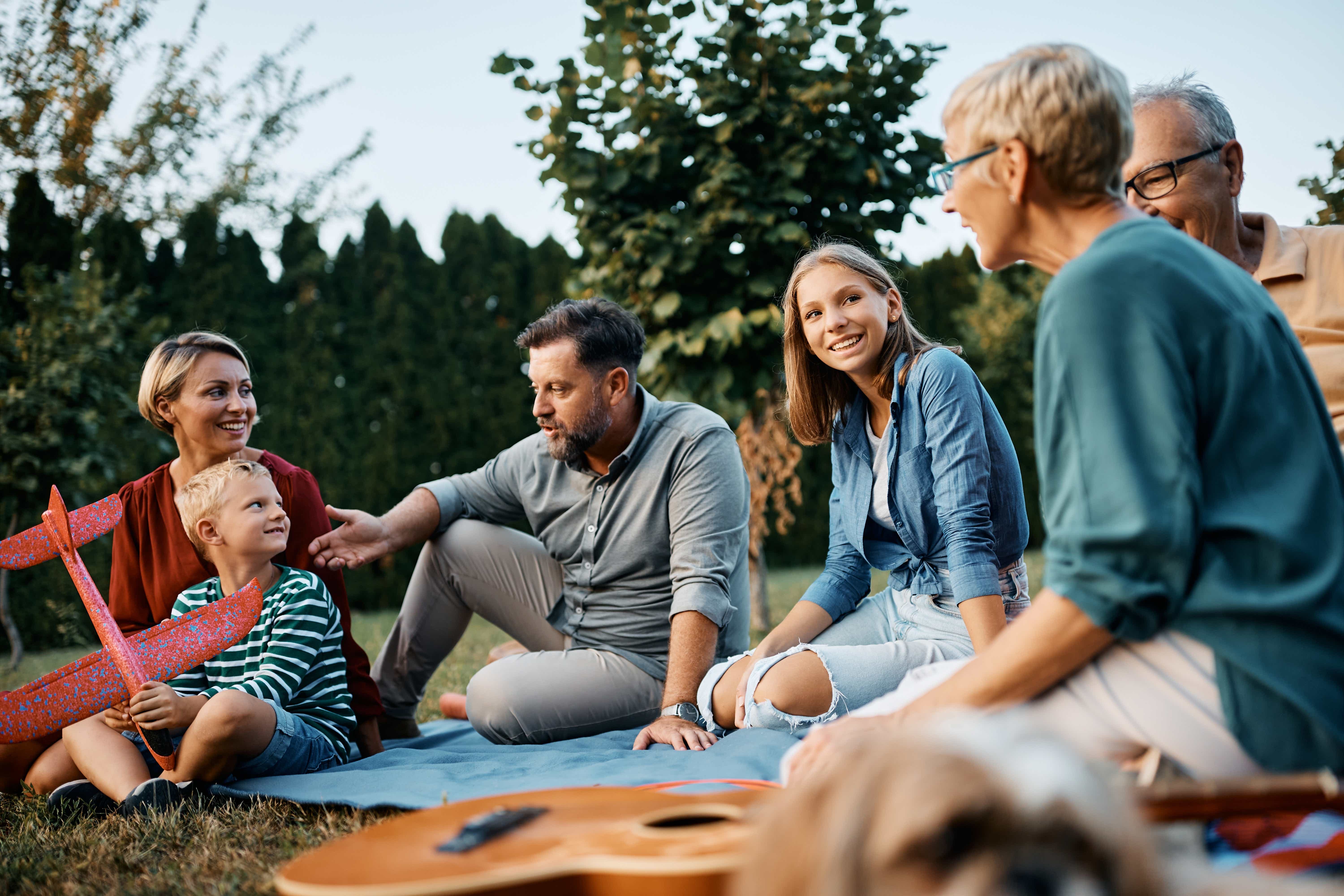 Mehrgenerationenfamilie sitzt auf einer Decke im Garten, lacht und unterhält sich. Ein Junge hält ein rotes Spielzeugflugzeug.