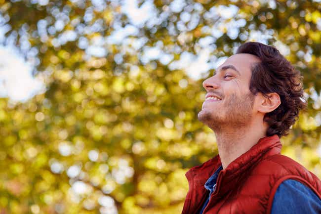 Ein Mann mittleren Alters steht in der Natur und hat seinen Kopf leicht in den Nacken gelegt. Er hat seine Augen geschlossen und lächelt zufrieden.