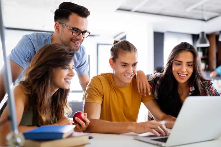 Eine Gruppe junger Leute blickt lächelnd auf einen auf einem Tisch stehenden Laptop. Drei der Personen sitzen, während ein Mann hinter ihnen steht.