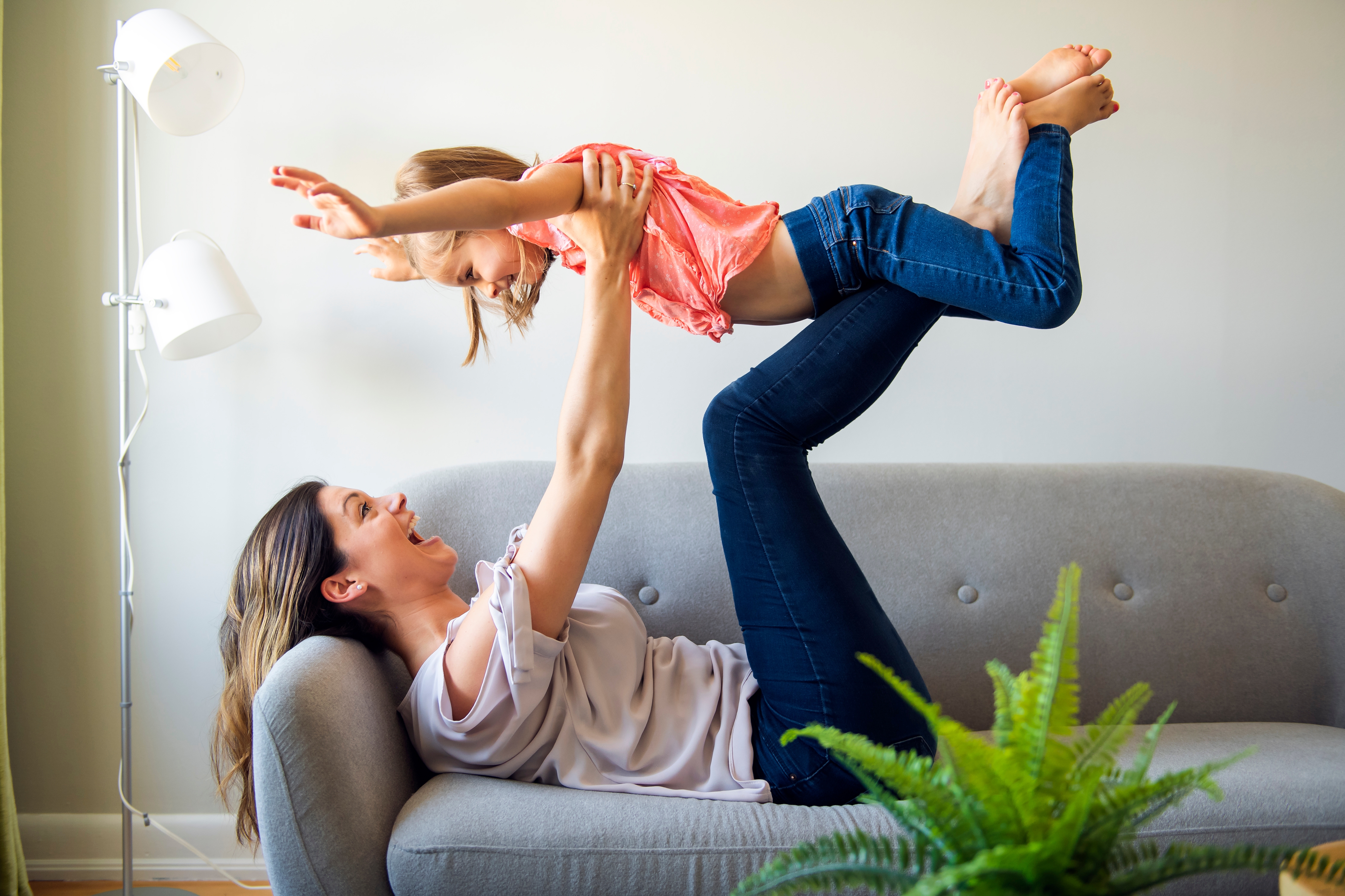 Lachende Mutter liegt auf dem Sofa und spielt mit ihrer Tochter, die sie mit den Beinen in die Luft hebt.