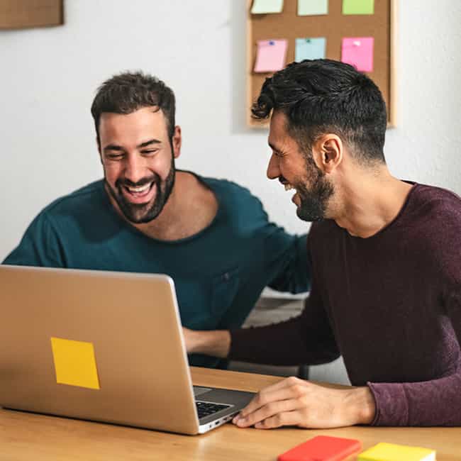 Zwei Männer mit dunklen Haaren sitzen an einem Tisch vor einem Laptop. Der eine Mann blickt glücklich auf den Bildschirm, während der andere lächelnd zu ihm sieht.