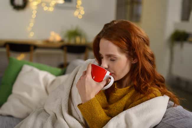 Eine Frau, in Strickpulli, liegt mit einer Decke eingewickelt auf einer Coach. Sie hält eine Tasse in der Hand und trinkt zufrieden davon.