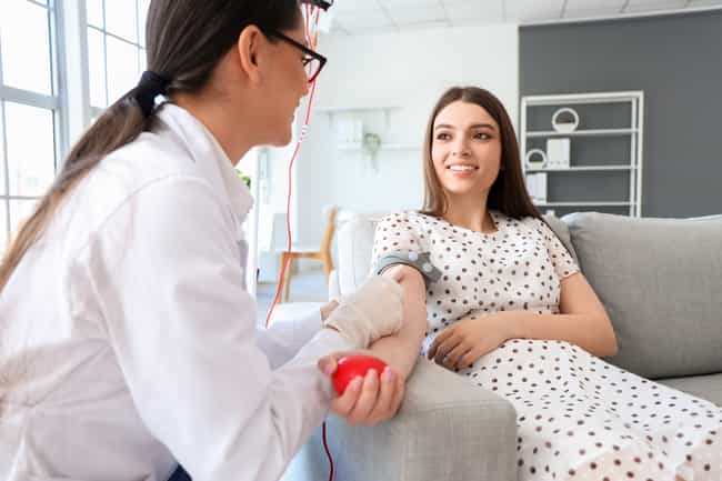 Junge Frau spendet Blut und lächelt eine Ärztin an, die ihr beim Ablauf hilft. Sie sitzt entspannt auf einem Sofa.