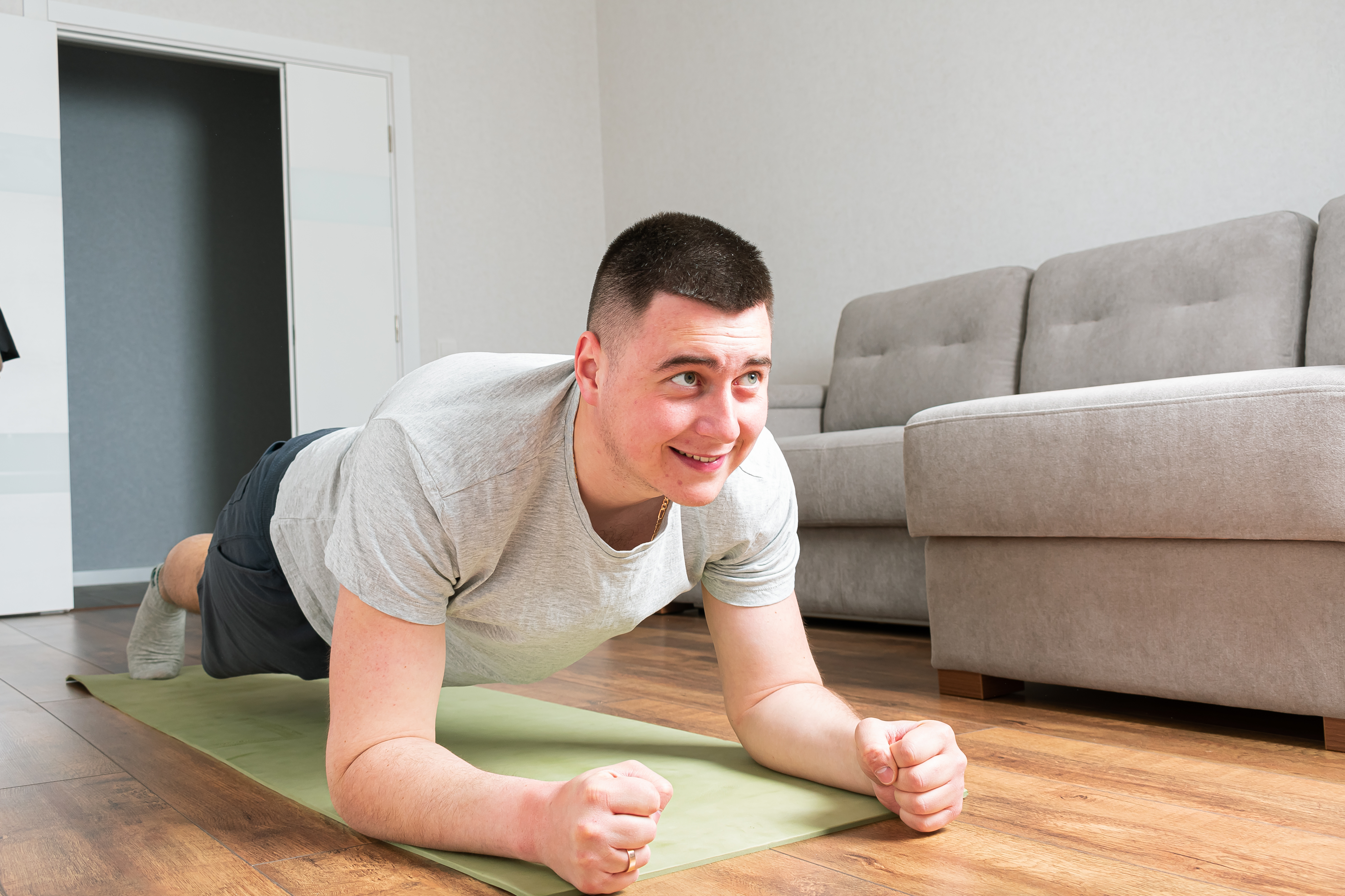 Ein junger Mann, in Sportkleidung, ist in Plank-Position auf einer Turnmatte im Wohnzimmer. 