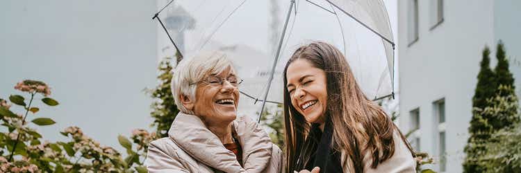 Eine junge und eine ältere Frau stehen unter einem Regenschirm und lachen gemeinsam.