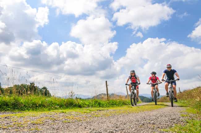 Drei Personen, mit Sportkleidung und einem Helm, fahren auf einem Feldweg Fahrrad. Die Sonne scheint und der Himmel ist blau.