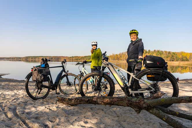 Zwei Personen, die einen Fahrradhelm tragen, stehen neben ihren Fahrrädern und stützen sich an ihnen ab. Im Hintergrund ist ein großer See.