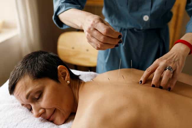 Frau mit kurzen Haaren erhält Akupunktur-Behandlung auf dem Rücken. Sie liegt entspannt auf einer Liege und lächelt.