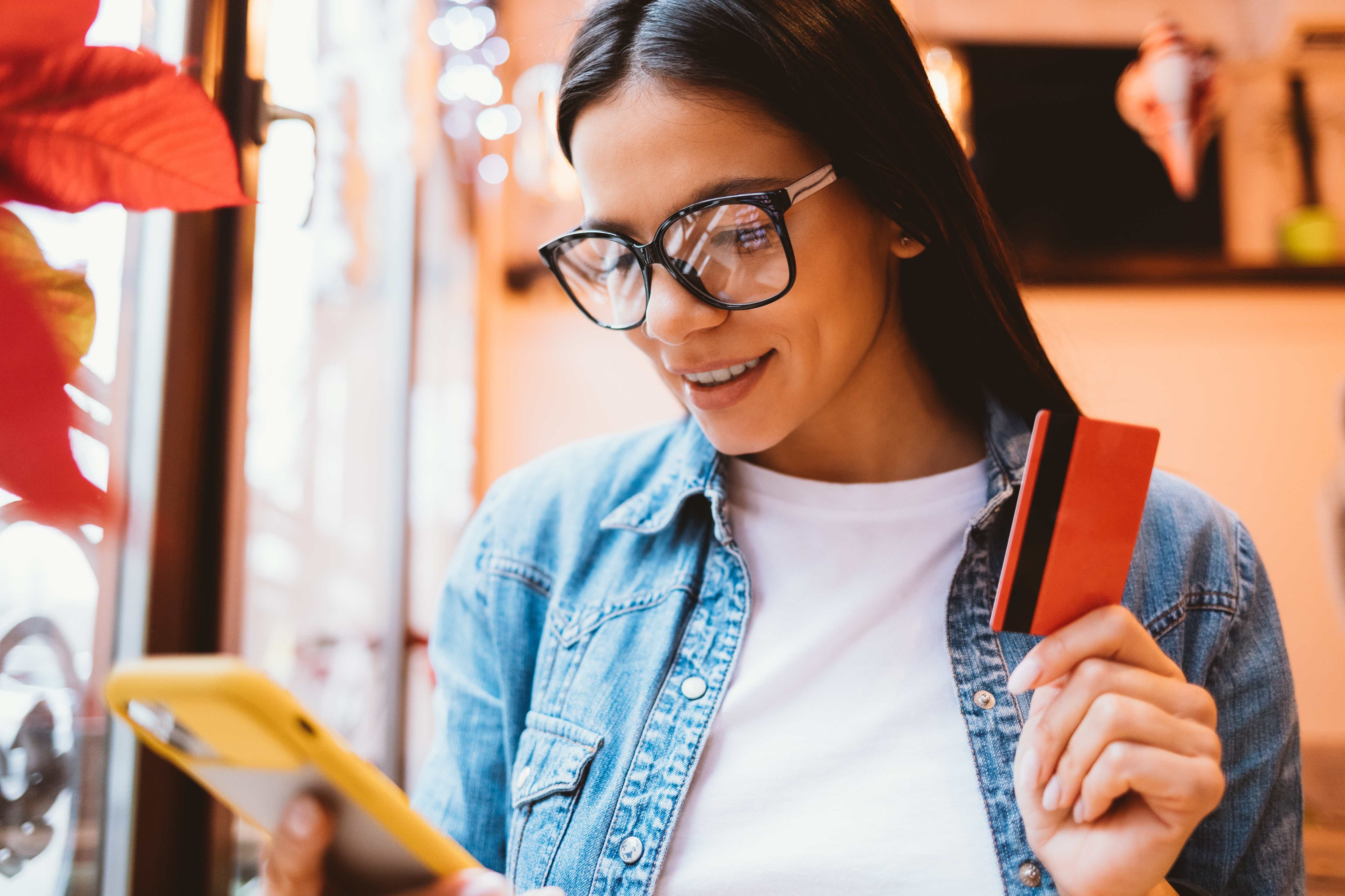 Junge Frau mit Brille hält eine Kreditkarte und schaut lächelnd auf ihr Smartphone.
