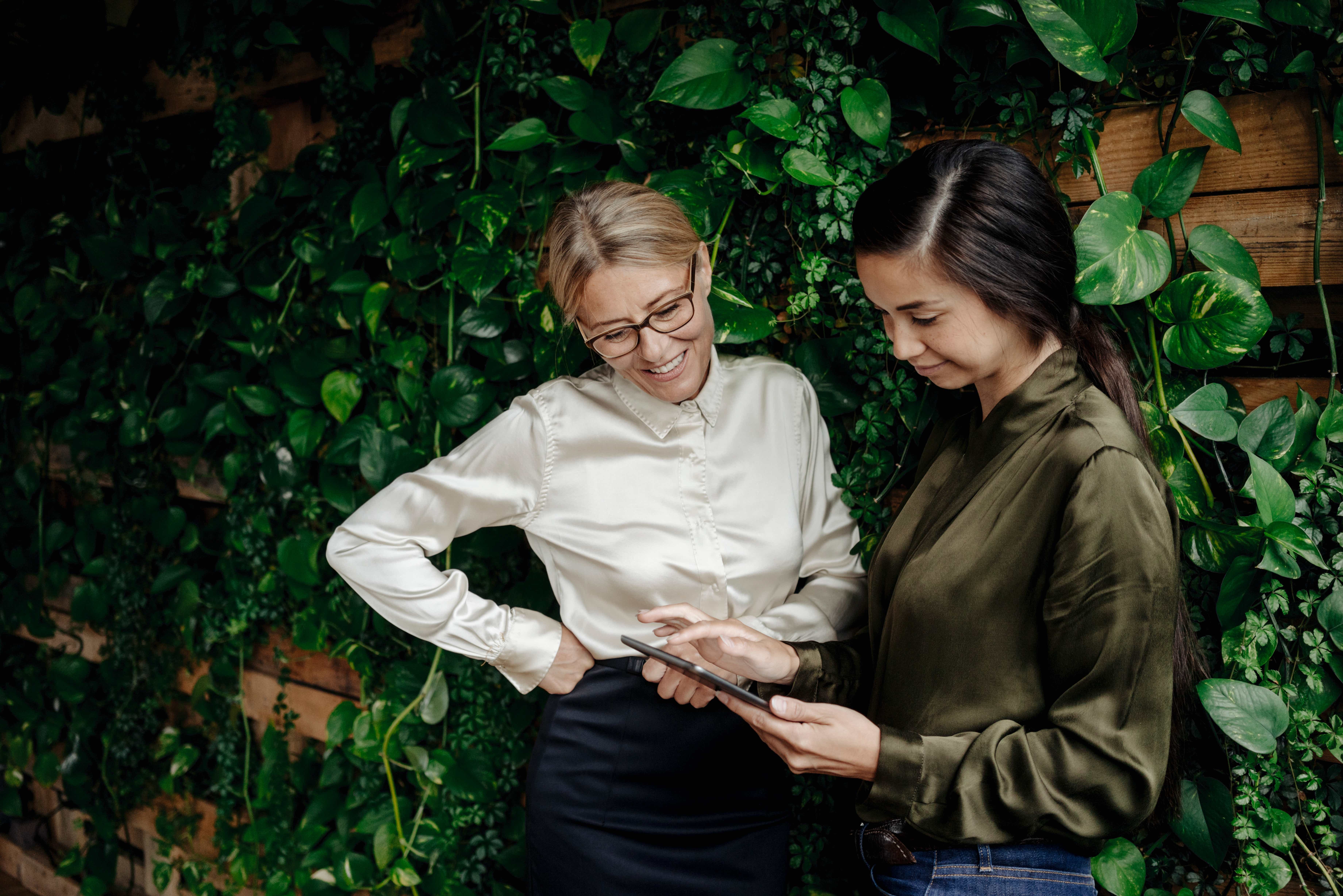 Zwei Frauen stehen vor einer grünen Pflanzenwand und schauen gemeinsam auf ein Tablet – sie wirken konzentriert und im Gespräch über nachhaltige Geschäftsideen.