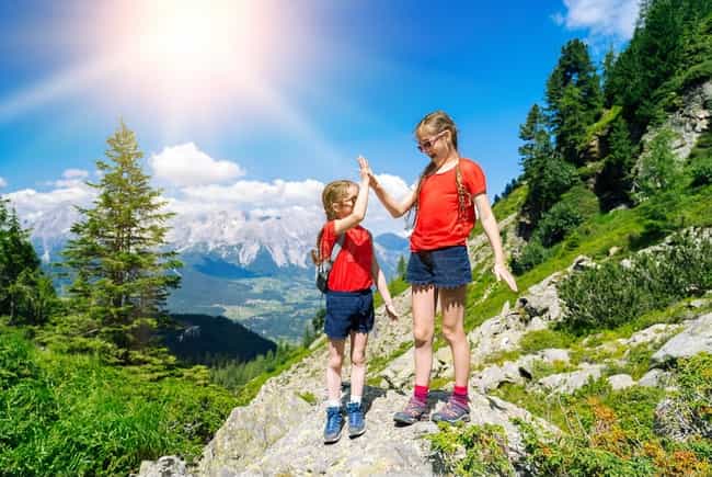 Zwei Mädchen in roten Shirts geben sich ein High-Five beim Wandern in den Alpen, mit Panoramablick auf Berge und sonnigem Himmel.