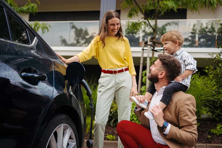 Eine Frau steht neben einem Elektroauto, welches gerade aufgeladen wird. Sie schaut auf einen Mann, der neben dem Auto hockt, und einen kleinen Jungen auf den Schultern trägt.