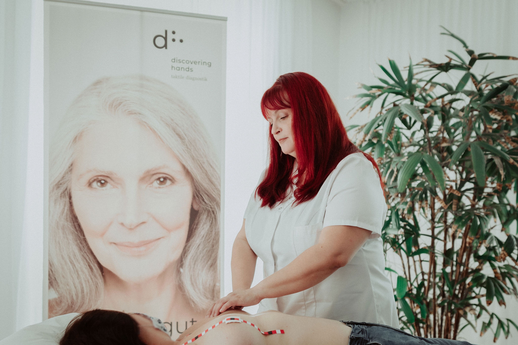 Eine Frau mit weißer Uniform führt eine Untersuchung an einer liegenden Patientin durch. Im Hintergrund ist ein großes Banner mit dem Logo „d: discovering hands“. 