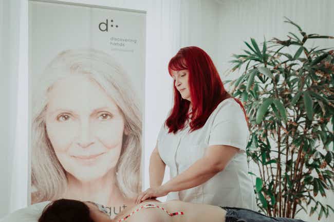 Eine Frau mit weißer Uniform führt eine Untersuchung an einer liegenden Patientin durch. Im Hintergrund ist ein großes Banner mit dem Logo „d: discovering hands“.