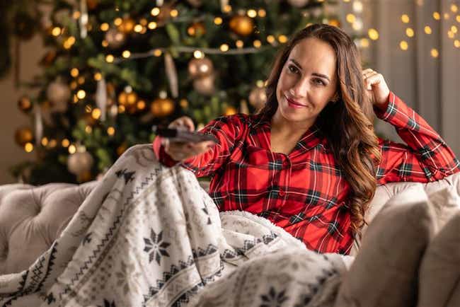 Person in rotem Karopyjama sitzt unter einer Decke und hält eine Fernbedienung, im Hintergrund ein geschmückter Weihnachtsbaum.