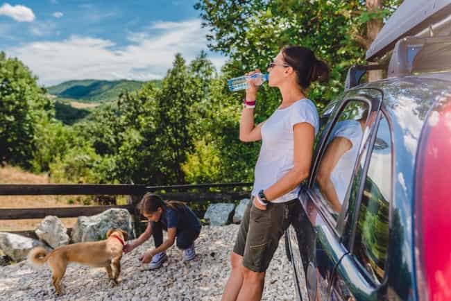 Eine Frau in Wanderkleidung, lehnt an einem Auto und trinkt vor saftig grüner Landschaft Wasser. Daneben streichelt ein Mädchen einen Hund.