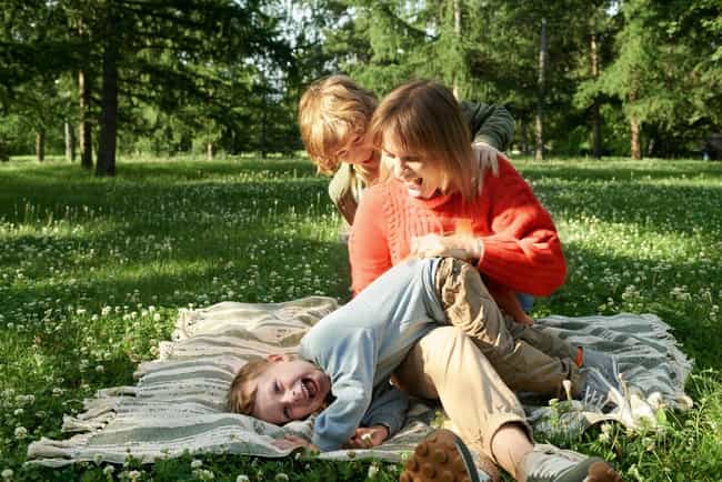 Mutter spielt lachend mit zwei Kindern auf einer Decke im Park. Sie genießen gemeinsam die Zeit in der Natur.