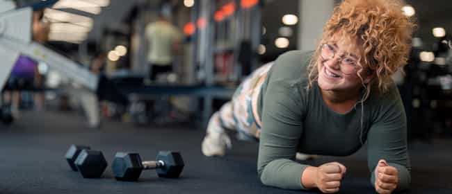 Eine Frau ist im Fitnesscenter, in Plank-Position. Sie ist sportlich bekleidet und neben ihr liegen Hanteln.