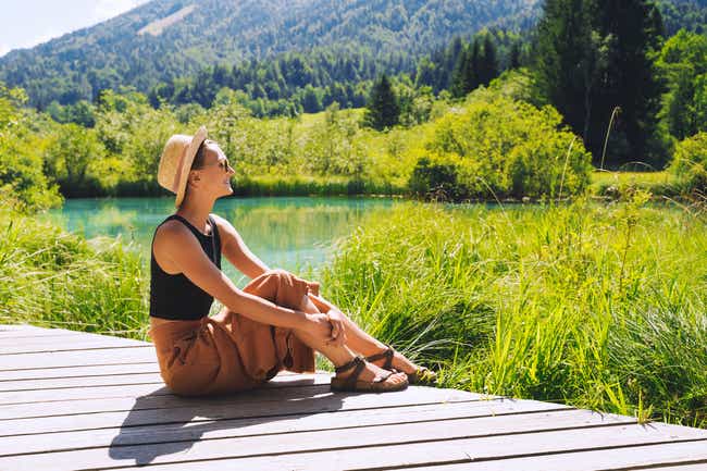 Eine junge Frau sitzt im Sonnenschein auf einem Holzsteg und blickt lächelnd in die Ferne. Umgeben ist sie von einem türkisen See und am Ufer sind viele grüne Gräser.