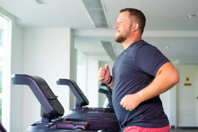 Ein festerer Mann läuft auf einem Laufband und schaut nach vorne. Das Bild wurde von der Seite gemacht.