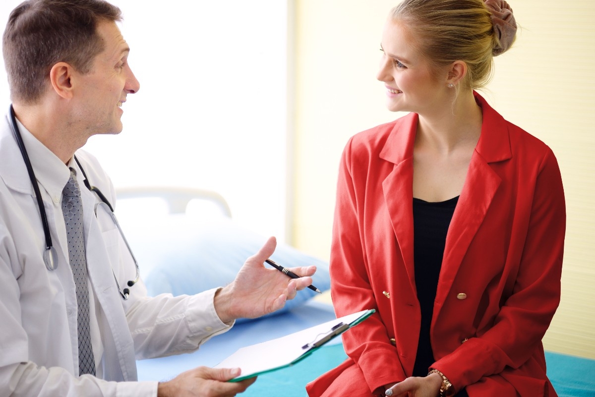 Eine junge Frau sitzt lächelnd neben einem Arzt mit weißem Kittel. Der Arzt hält einen Zettel in seiner Hand und erklärt der Frau etwas.