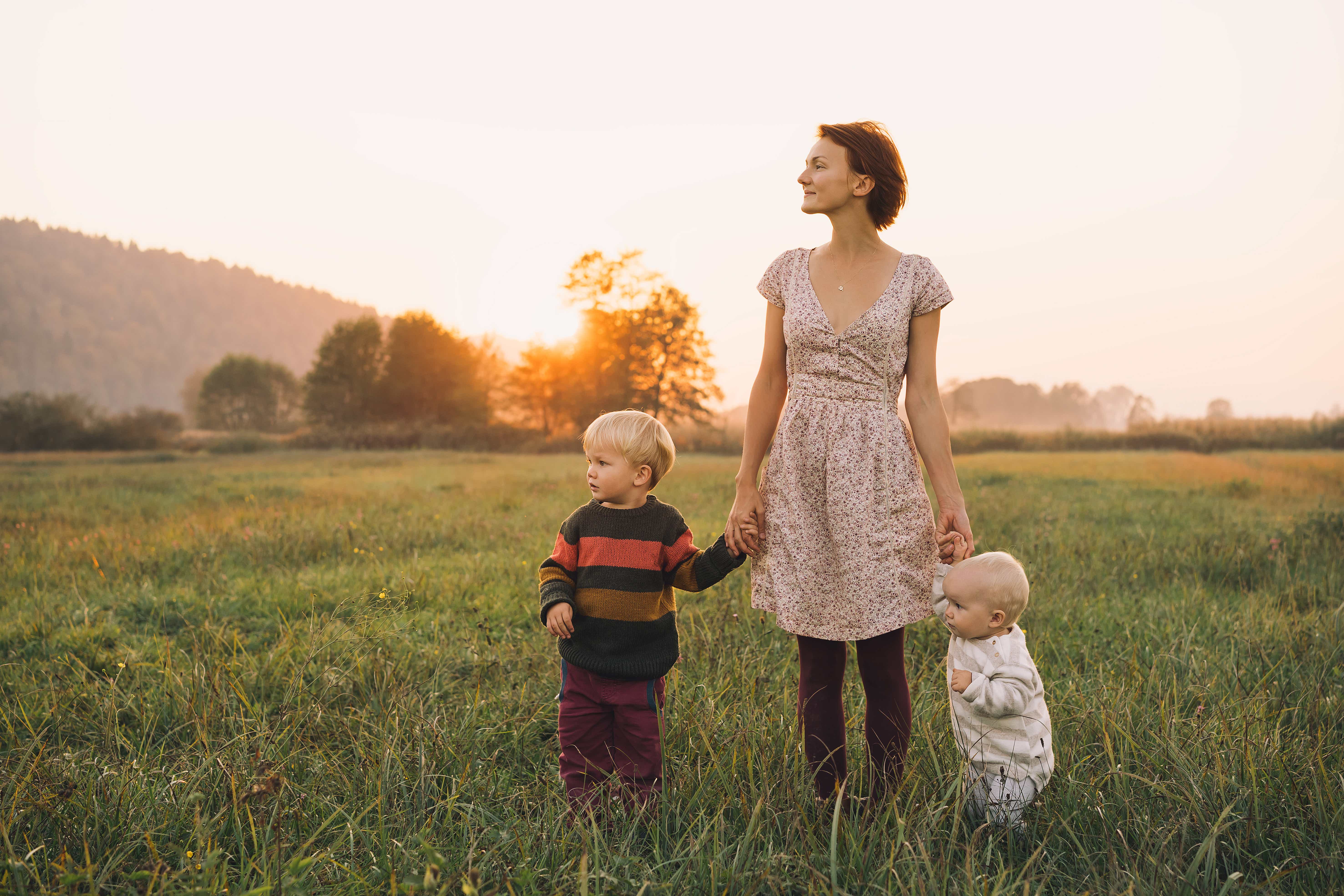 Eine Frau in einem Blumenkleid steht beim Sonnenuntergang auf einer Wiese und blickt in die Ferne. An ihren beiden Händen halten sich, links und rechts, je ein Kleinkind fest. 