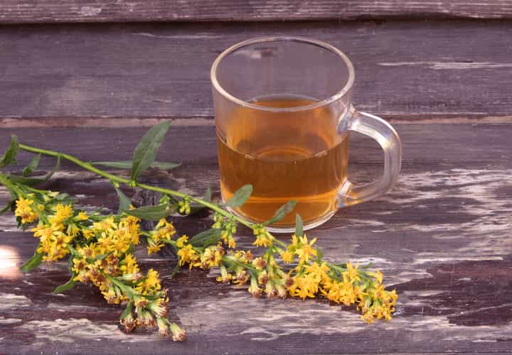 Ein Glas mit Henkel, welches mit Tee befüllt ist, steht auf einem Holztisch. Daneben liegen Kräuter.