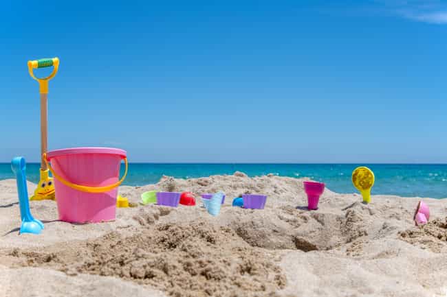 An einem sonnigen Sandstrand befindet sich verschiedenstes Sandspielzeug, wie eine Schaufel und ein Eimer. Im Hintergrund fließt klares, blaues Meerwasser.