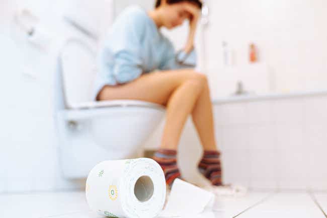 Eine Frau sitzt auf der Toilette im Badezimmer und fasst sich mit der Hand an die Stirn. Im Vordergrund liegt eine Rolle Toilettenpapier auf dem Boden.