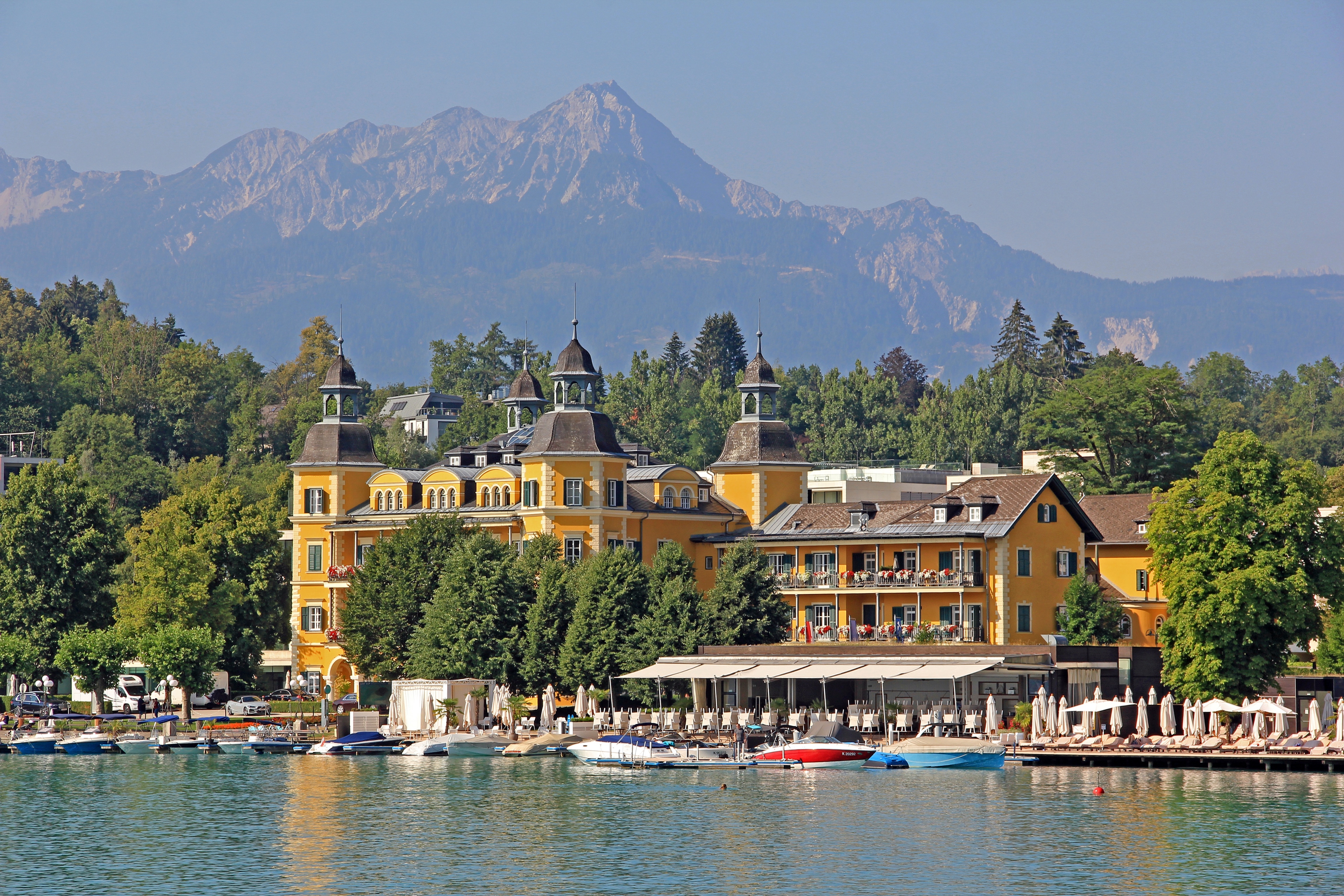 Ein gelbes, großes Hotel steht am Ufer des Wörthersees. Im Hintergrund sind Berge. 