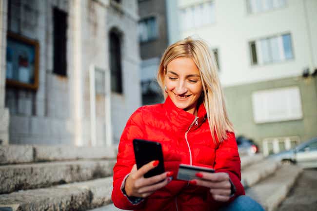 Eine Frau mit roter Jacke blickt lächelnd auf ein Handy, welches sie in ihrer Hand hält. Im Hintergrund befinden sich Häuser.