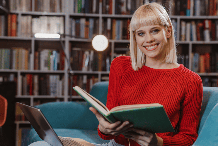 Lächelnde junge Frau mit blondem Bob und rotem Pullover sitzt in einer Bibliothek, liest ein Buch und nutzt einen Laptop.