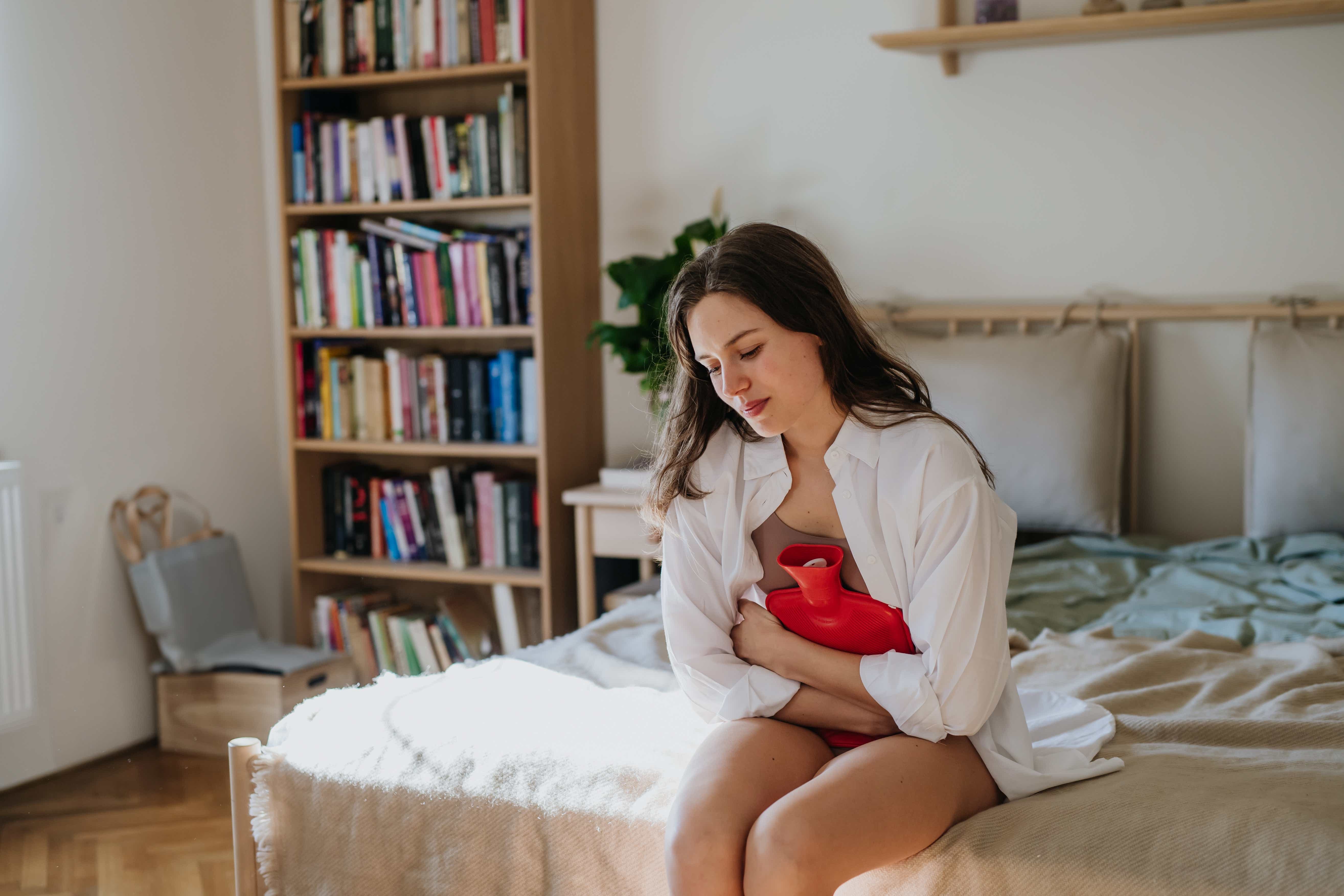 Junge Frau sitzt mit Wärmflasche auf dem Bett, hält sich den Bauch und wirkt nachdenklich. Im Hintergrund steht ein Bücherregal.